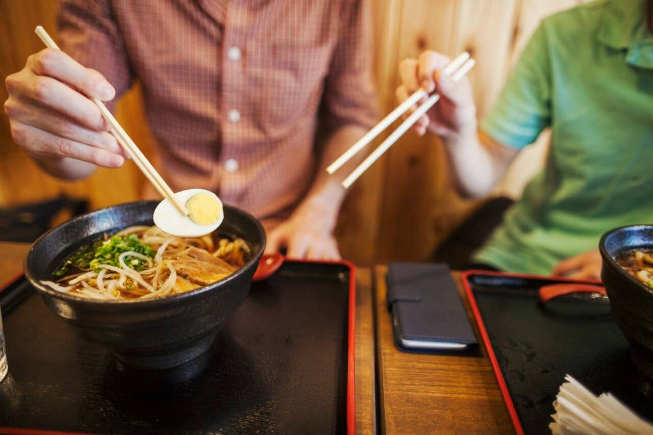 Two people, a Japanese man showing a Western man how to use chopsticks in a noodle shop.